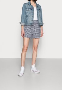 Persona che indossa una giacca di jeans arrotolata, una maglietta bianca, pantaloni corti a quadretti blu e bianchi e sneaker bianche alte, in piedi su un pavimento grigio.