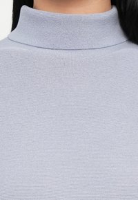 Izbliza prikaz svijetlosive pletene turtleneck džemperice na osobi s dugom tamnom kosom.