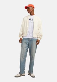 Lys beige bomberjakke over en hvid grafisk t-shirt, blå afslappede jeans og grå sneakers med sorte stropper. En orange kasket fuldender looket.