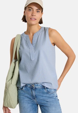 Femme portant un chemisier sans manches à rayures bleues, un jean, une casquette beige et un sac matelassé vert clair à l'épaule.