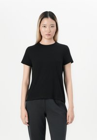 Jeune femme aux cheveux raides portant un t-shirt noir uni à manches courtes et un pantalon sombre, se tenant devant un fond blanc.