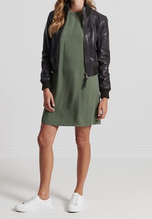 Femme portant une robe courte vert olive, une veste aviateur en cuir noir, des baskets blanches et un collier chaîne en or sur un fond uni.