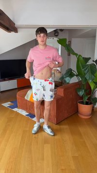Jauns vīrietis paceļ rozā kreklu, lai parādītu vēderu, valkā baltas rakstainas šortus un sudraba sporta apavus, stāv dzīvojamā istabā ar augiem un dīvānu.