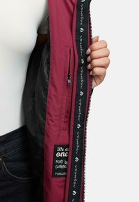 Czerwono-brązowa kurtka wewnętrzna z czarną, fakturowaną podszewką. Wyposażona w kieszeń na zamek błyskawiczny oraz metkę z napisem "Jesteśmy jednością" w białym kolorze.