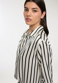 Blusa bianca con strisce verticali nere; presenta un colletto, maniche lunghe e un leggero scollo a V; texture liscia.