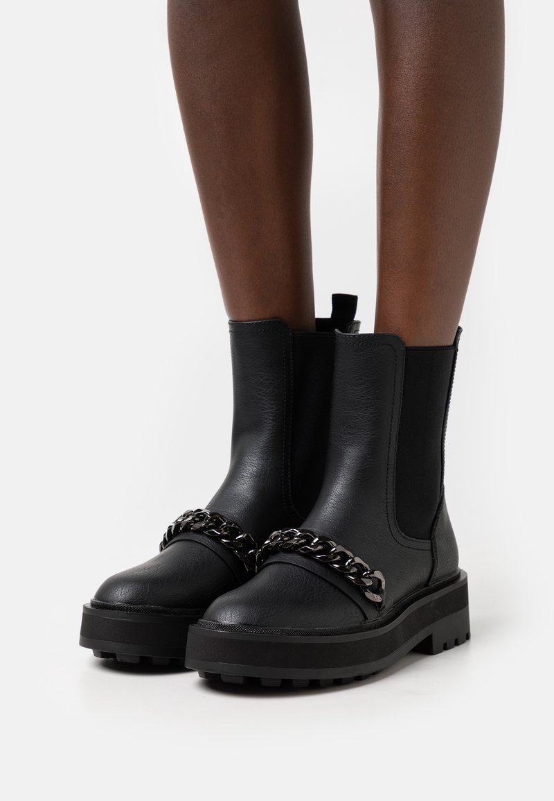 Bottes cheville en cuir noir avec semelles épaisses et détail chaîne argentée portées sur des jambes à peau foncée sur fond blanc.