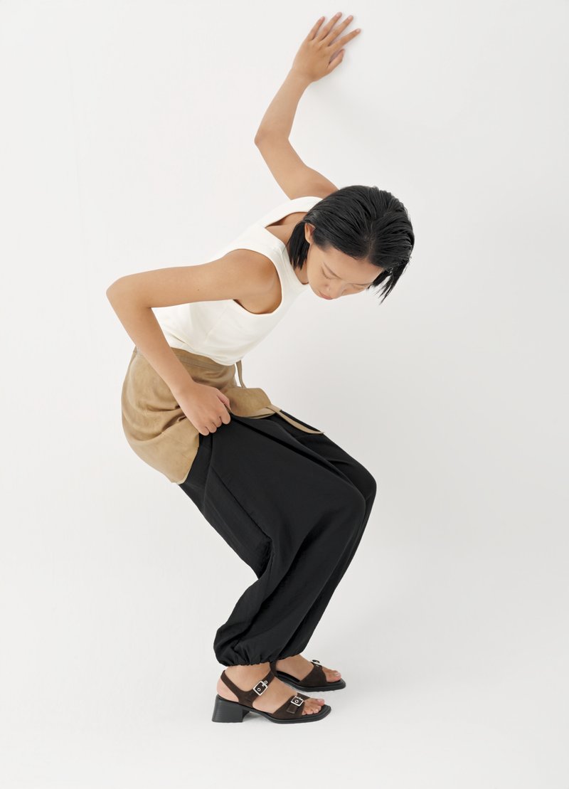 Frau in weißem, ärmellosem Oberteil, brauner Schürze und schwarzer Hose, die sich nach vorne beugt und mit einer Hand eine weiße Wand berührt.