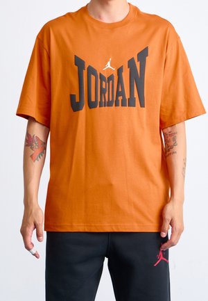 Person trägt ein oranges Jordan-T-Shirt mit schwarzer Schrift und schwarze Hosen mit rotem Jordan-Logo, sichtbare Tätowierungen am Arm und bandagierter Finger.