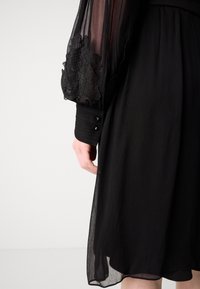 Robe noire avec des manches longues transparentes ornées de dentelle noire, tissu texturé et détails de boutons au niveau des poignets. Jupe fluide et lisse.