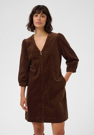 Femme portant une robe marron en velours côtelé à manches trois-quarts et une fermeture éclair frontale, debout avec une main dans sa poche.