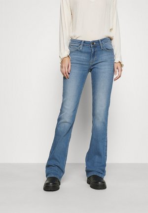 Person iført lyseblå flared jeans, cremefarvet bluse med lange ærmer og sorte chunky støvler står foran en ensfarvet baggrund.