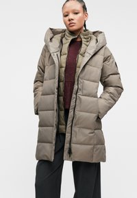 Pufová bunda svetlohnedej farby s kapucňou, obsahujúca prešívané panely, predné gombíky a vrstvenú vnútornú vestu olivovej zelenej farby.