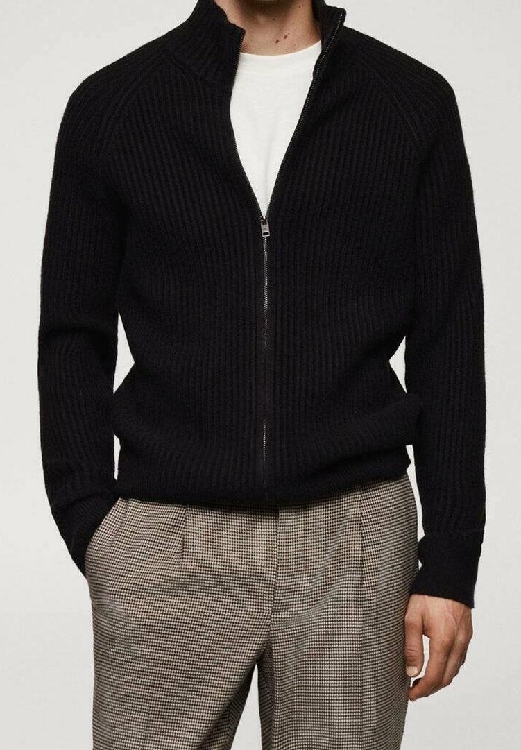 Personne portant un pull noir côtelé zippé sur une chemise blanche et un pantalon beige à carreaux, les mains dans les poches.