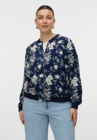 Navy floral bomber jacket with a zipper, featuring cream flowers and green accents, elastic cuffs, and a relaxed fit. Paired with light jeans.
