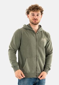 Sweat à capuche zippé vert en tissu doux, avec des poches avant, des poignets côtelés et une capuche avec cordon de serrage. Un logo est visible sur la poitrine.