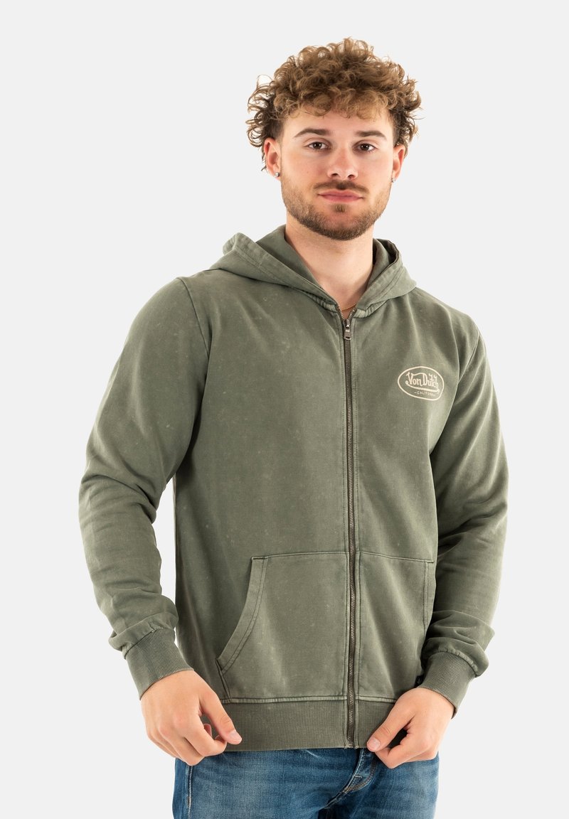 Sweat à capuche zippé vert en tissu doux, avec des poches avant, des poignets côtelés et une capuche avec cordon de serrage. Un logo est visible sur la poitrine.