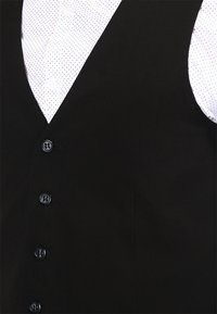 Schwarze Weste mit glatter Textur und sechs vorderen Knöpfen. Getragen über einem weißen Hemd mit einem kleinen schwarzen Punktmuster.