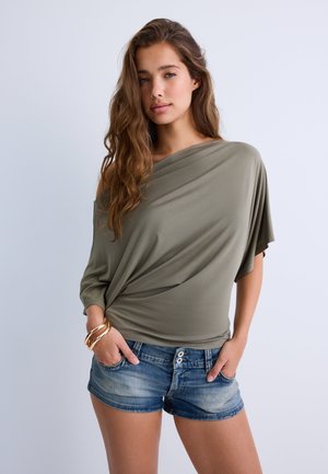 Junge Frau mit langem welligem Haar, die ein olivgrünes schulterfreies Oberteil, Jeansshorts und goldene Armbänder trägt und mit den Händen in den Taschen steht.