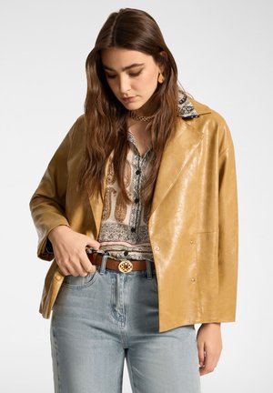 Donna con i capelli castani lunghi che indossa una giacca di pelle color cuoio, una camicetta con motivo paisley, una collana a catena dorata e jeans azzurri, mentre sistema la cintura marrone.