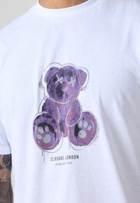 Hvit bomull t-skjorte med et lilla bjørnegraphic med X-formede øyne og poter. Logo teksten "CLOSURE LONDON" under designet.