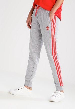 Osoba nosząca szare spodnie dresowe Adidas z czerwonymi paskami i logo, w połączeniu z białymi butami sportowymi na rzepy.