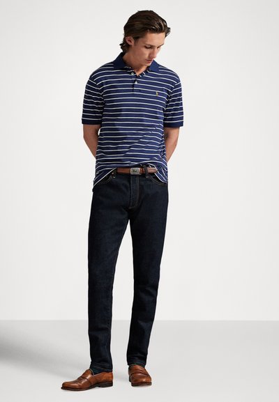 Jauns vīrietis stāv ar rokām aiz muguras, valkā tumši zilas svītras polo kreklu, tumšas džinsas, brūnu jostu un brūnas loafer kurpes uz vienkārša fona.