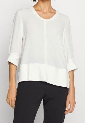 Femme portant un haut blanc ample à manches trois-quarts avec un large ourlet, associé à un pantalon noir ajusté.