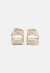 Beige sandaler har et bredt stroppdesign, åpen hæl og strukturert såle. Merkenavnet er preget på hælen.