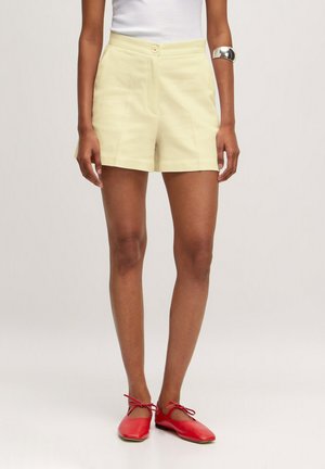 Persoon draagt lichtgele shorts met hoge taille, wit mouwloos topje, zilveren manchetarmband en rode balletschoenen met enkelbandjes op een effen achtergrond.