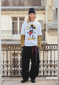 Bijela majica s grafikom Mickey Mouse, žutim natpisom i crnim prugama na rukavima; uparena s tamnim hlačama i crvenom maramom.