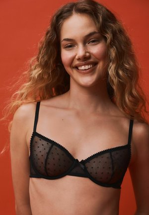 Young woman with long curly hair smiling, wearing a black sheer polka dot bra against a solid orange background.