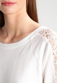 Indumento bianco con una texture morbida e dettaglio in pizzo sulla spalla. Il colletto è ampio e le maniche sono lunghe.