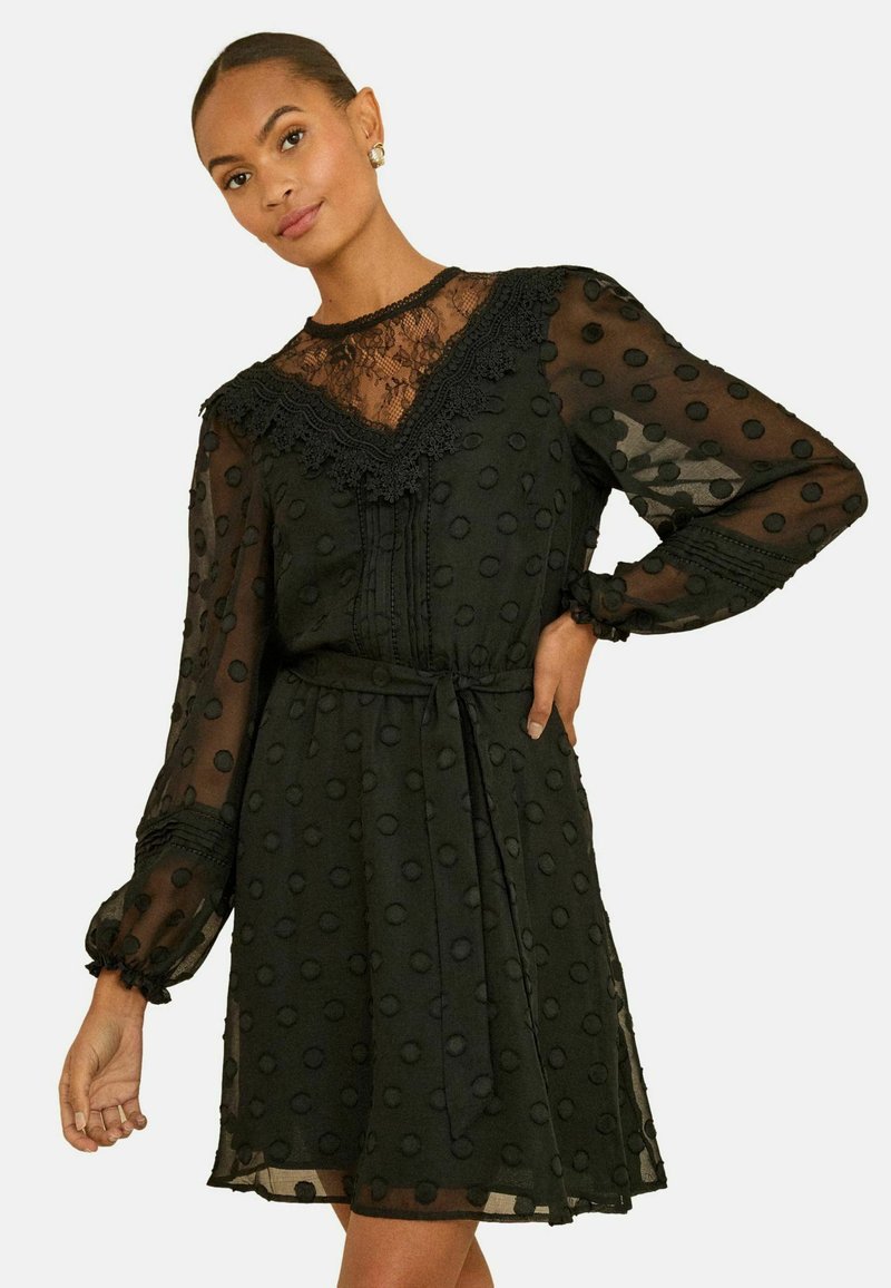 Rochie neagră din material transparent, cu buline texturate. Prezintă un guler din dantelă, mâneci lungi și un detaliu cu fundă la talie.