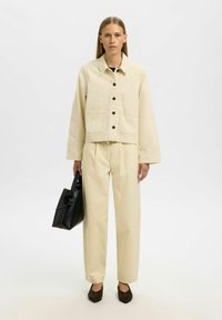 Kremowa krótka kurtka z czarnymi guzikami i dwoma przednimi kieszeniami; pasujące, wysokie spodnie; trzyma czarną torebkę; ciemne buty.