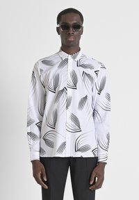 Camisa blanca de botonadura con patrones negros en forma de hoja, mangas largas y un cuello clásico. Llevada con pantalones negros y gafas de sol.
