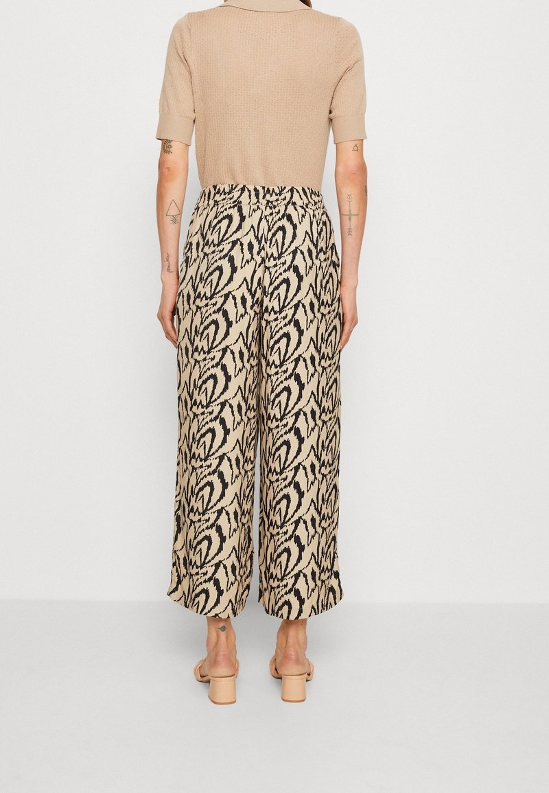 Femme debout de dos, portant un haut beige à manches courtes, un pantalon beige à larges jambes avec un imprimé abstrait noir, et des sandales à talons beiges.