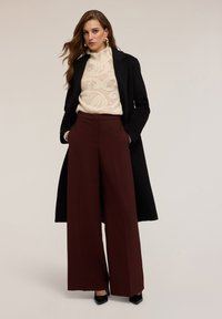 Žena v černém dlouhém kabátu, krémovém pleteném svetru, širokých hnědých kalhotách a černých špičatých lodičkách, stojící se založenýma rukama v kapsách na jednobarevném pozadí.