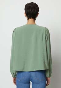 Blusa verde a maniche lunghe con una vestibilità morbida, scollatura rotonda e cuciture sulle spalle arricciate. Indossata con jeans blu che mostrano una leggera elasticità.