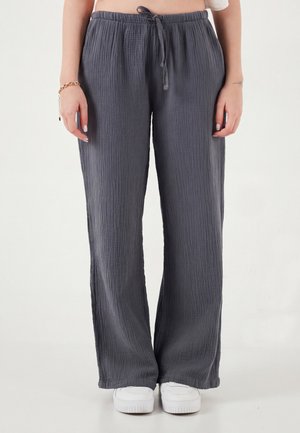 Kobieta nosząca luźne, ciemnoszare pogniecione spodnie z elastycznym pasem i sznurkiem, w połączeniu z białymi trampkami na jasnym tle.