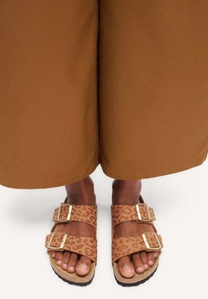 Voeten met tan luipaardprint sandalen met gouden gespen, gecombineerd met brede bruine broeken die boven de enkels zijn afgeknipt.