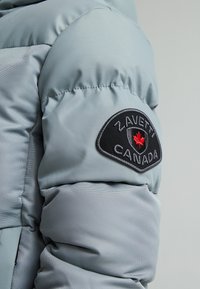 Doudoune gris clair avec un motif matelassé. Présente un patch noir avec une feuille d'érable rouge et le nom de la marque "Zavetti Canada" sur la manche.