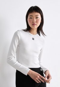 White long-sleeve top with round neckline, featuring "HUGO" logo in black on the chest, paired with black fitted bottoms. Smooth texture.
