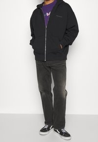 Person som bär en svart zip-up hoodie, lila tröja med fjärilsdesign, urblekta svarta jeans och svart-vita gympaskor.