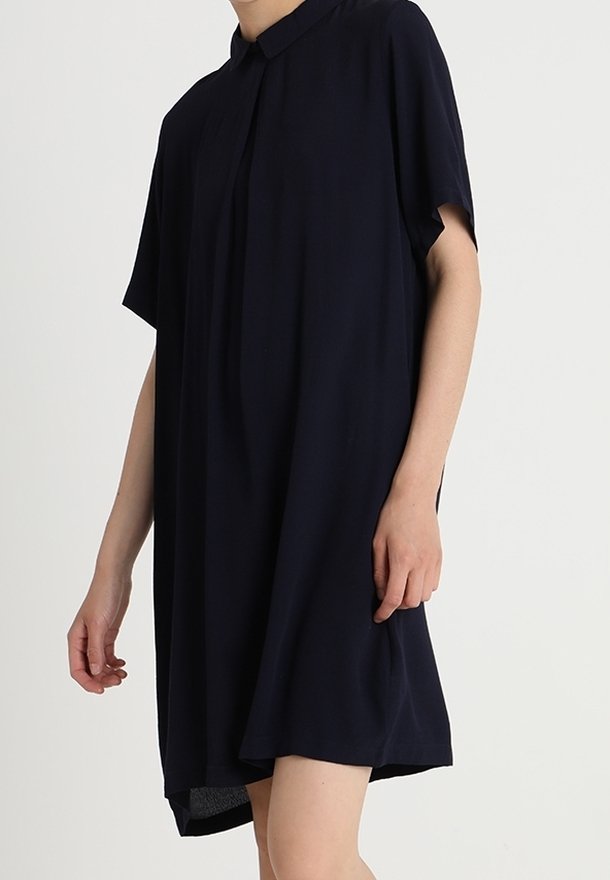 Femme portant une robe noire ample, à manches courtes, longueur genoux, avec un col et des boutons cachés à l'avant.