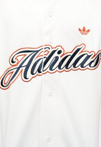 Biała koszula zapinana na guziki, z dużym granatowo-pomarańczowym napisem stylizowanym na pismo odręczne "Adidas" oraz małym pomarańczowym logo trefoil Adidas w pobliżu kołnierzyka.