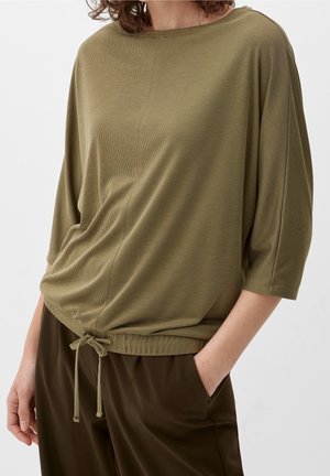 Femme portant un haut ample à manches longues vert olive avec ourlet à cordon de serrage et un pantalon brun foncé, une main dans la poche, sur fond uni.