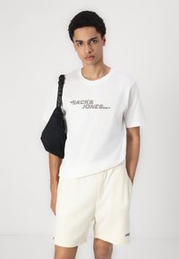 Camiseta blanca con gráfico de "JACK & JONES" en gris. Pantalones cortos color crema. Mochila cruzada negra con correa con logo. Mangas cortas, ajuste casual.
