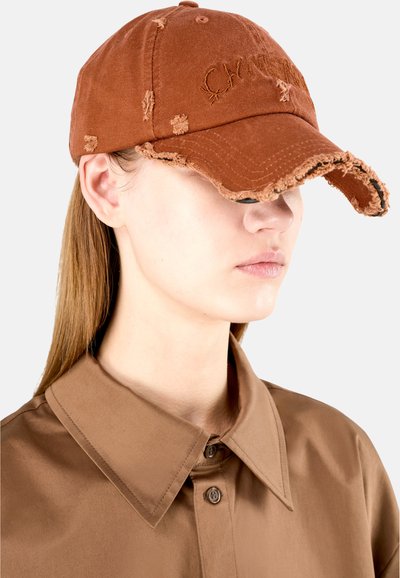 Gorra de béisbol marrón desgastada con bordes deshilachados y logo bordado. Combinada con una camisa de cuello marrón, la tela parece suave.