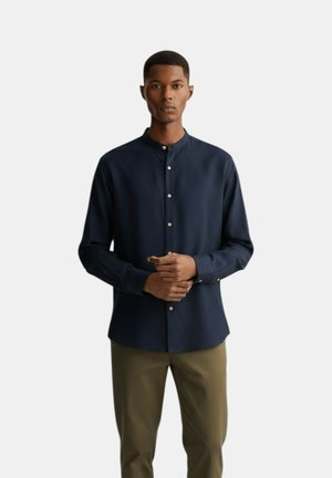 Uomo che indossa una camicia blu scuro abbottonata con colletto alla coreana e pantaloni verde oliva, in piedi con le mani giunte davanti.
