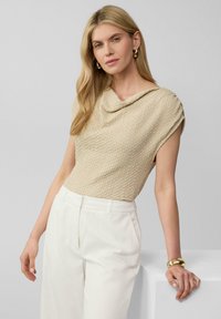 Beige textuurtop met een gedrapeerde kraag en kapmouwen, gecombineerd met witte, op maat gemaakte broeken. Gouden manchetarmband om de pols van het model.